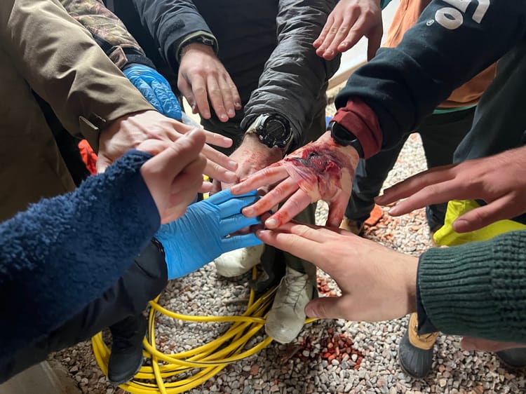 A set of hands meeting in the center of a circle—some gloved, some covered in mock blood—right before a "Go team!" chant