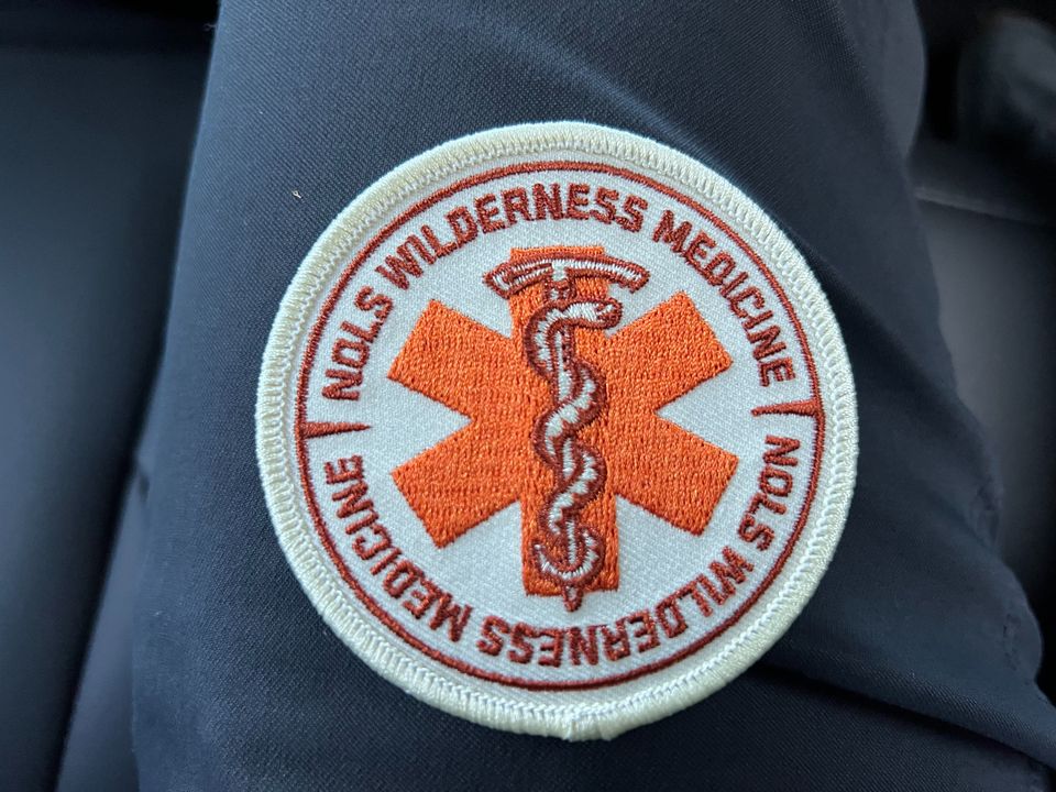 A white/orange patch, with a six-sided medical star and a snake wrapped around an ice axe, text: NOLS Wilderness Medicine